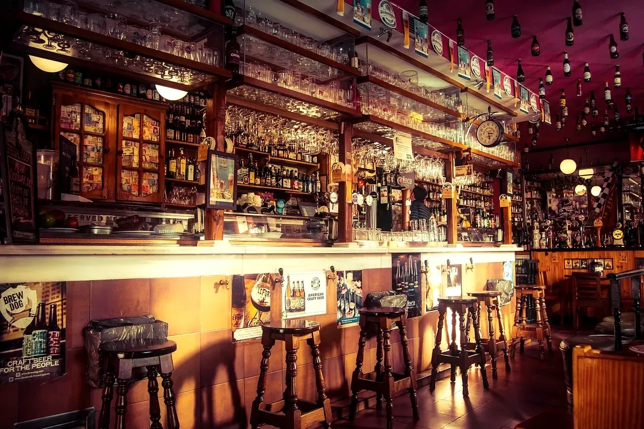 Warm-lit bar counter with a row of wooden stools and shelves of glassware